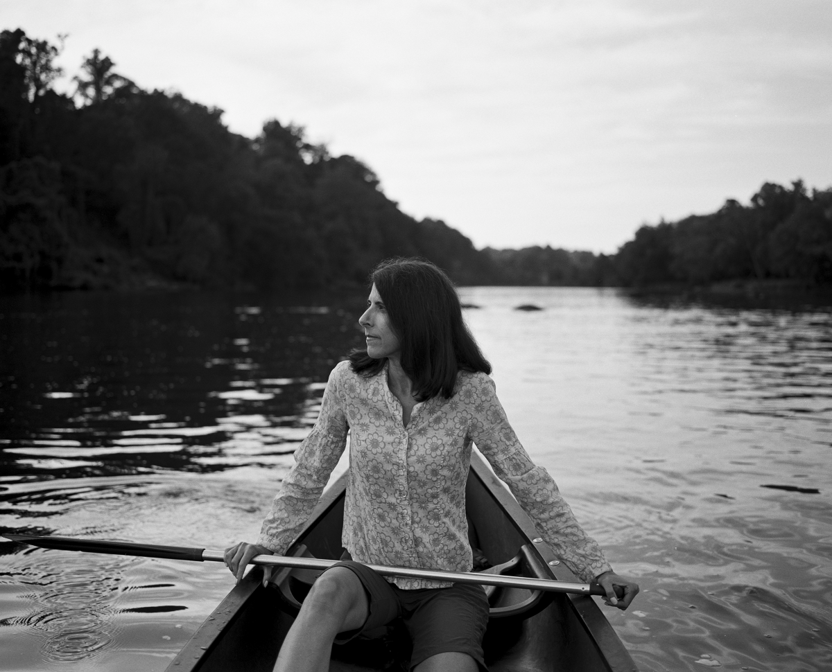 A black and white photo of a woman, Florence Williams, an Arc'tyrex ambassador, sitting in a canoe on the water, looking to her right.