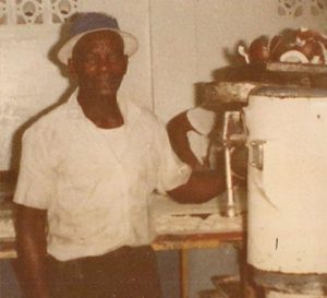 Old film photo of Charles Neale, a black man wearing a white button up shit and a blue brimmed hat, standing next to an ice cream machine.