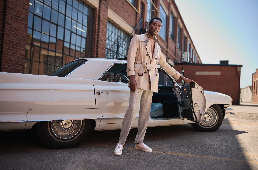 Mean standing beside white car against factory window backdrop looking confident wearing beige safari jacket and opening door