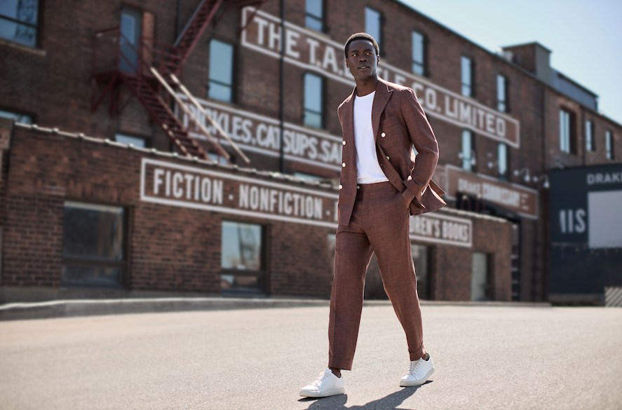 Man walking confidently against industrial backdrop wearing white t-shirt and brown linen suit with white sneakers