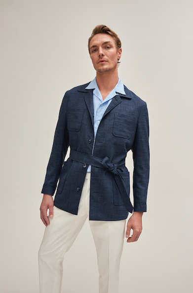 Man against neutral background wearing navy safari jacket with light blue shirt underneath and creme trousers