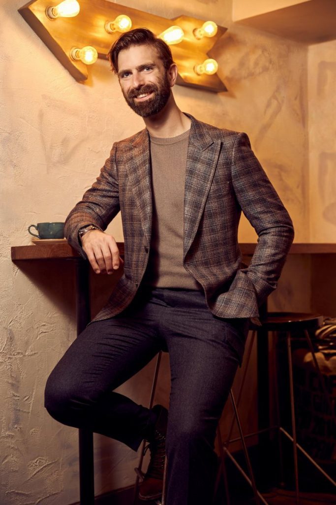 Ian Rosen in sports jacket, sweater and pants leaning against coffee table