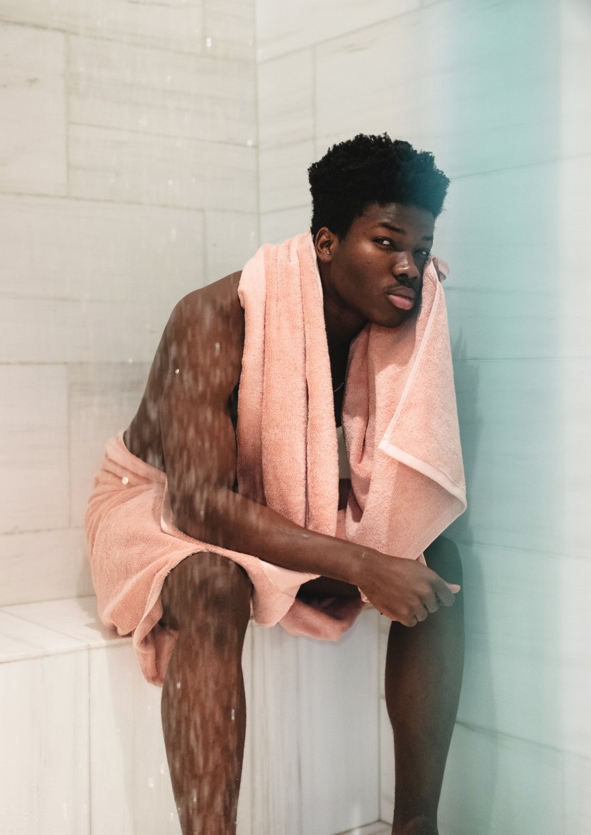 A man sits in the shower wearing a pink towel from Aestate.