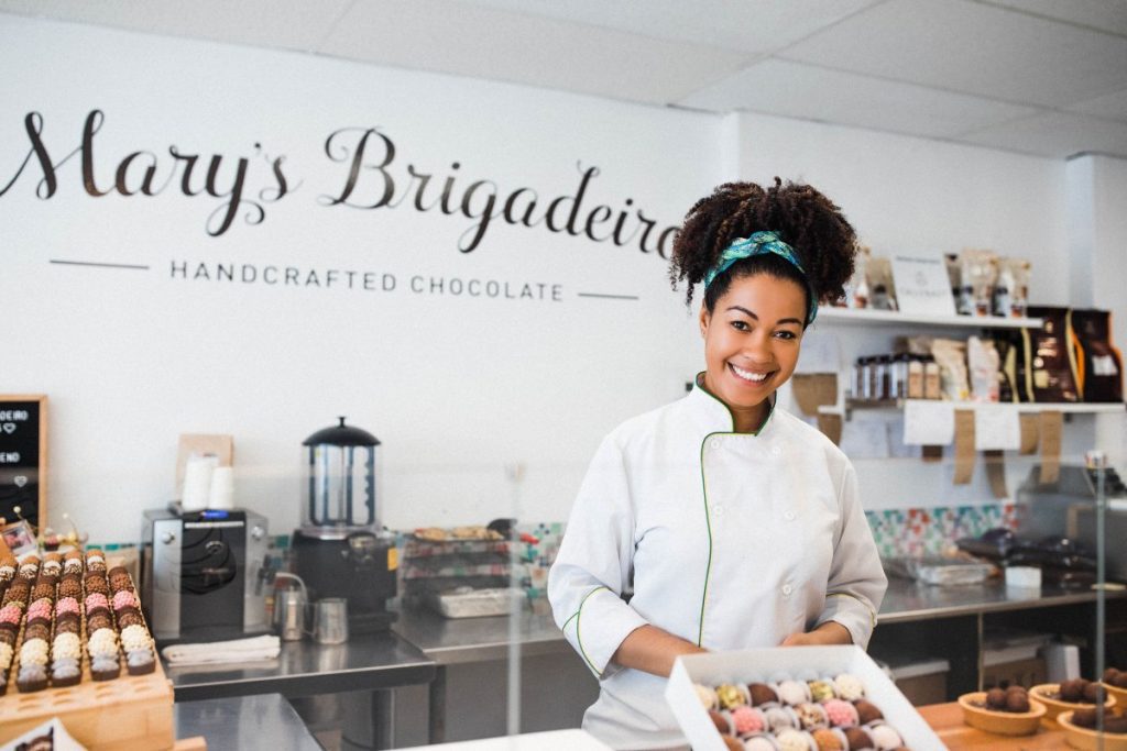 Woman in a white chef's jacket holds a tray of small round chocolates in a kitchen, to represent how the Black Entrepreneur Startup Program wants to help young Black entrepreneurs succeed.