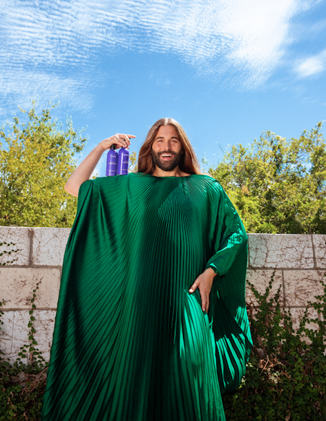 Jonathan Van Ness in pleated green caftan with beauty products on shoulder against nature backdrop