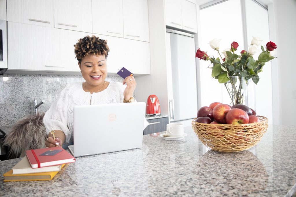 Vanessa Bowen, Creator of Mint Worthy at her Kitcen counter with her laptop, holding up a credit card. Her countertop has a vase of flowers and a fruit basket on it.