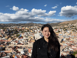 An asian woman, co-founder of Deel, Shuo Wang, stands in front of a city landscape.