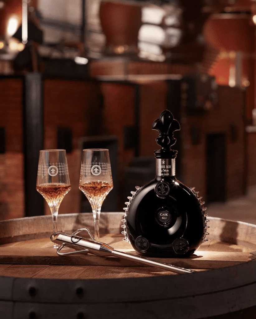 A black glass decanter of Louis XIII Rare Cask 42.1 cognac with two crystal glasses filled with cognac. The setting is in a old-fashioned bar.