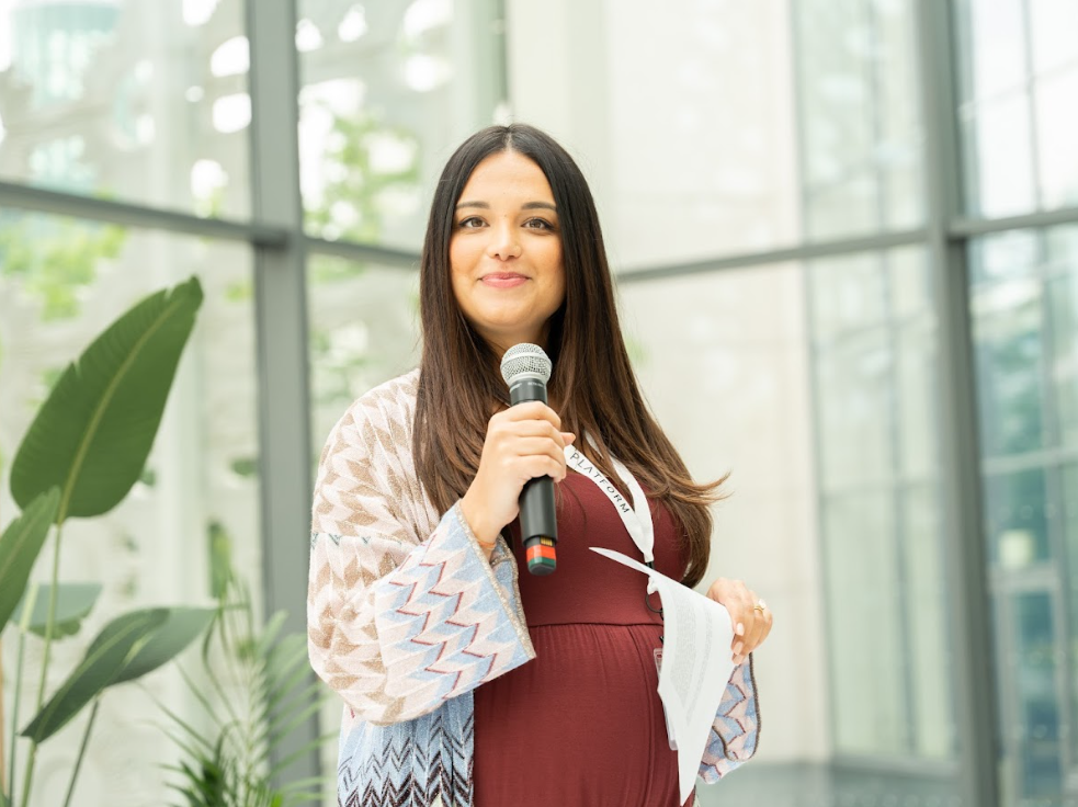Platform Social founder Sarah Koonar wearing light textured cardigan with burgundy blouse underneath. Sarah is holding a microphone and against a backdrop with windows and plants.
