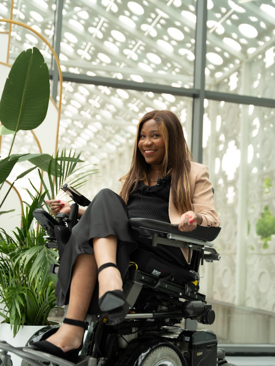 Taylor Lindsay-Noel, wearing all black with a beige blazer, is sitting in front of a wall of glass panels with plants around her.