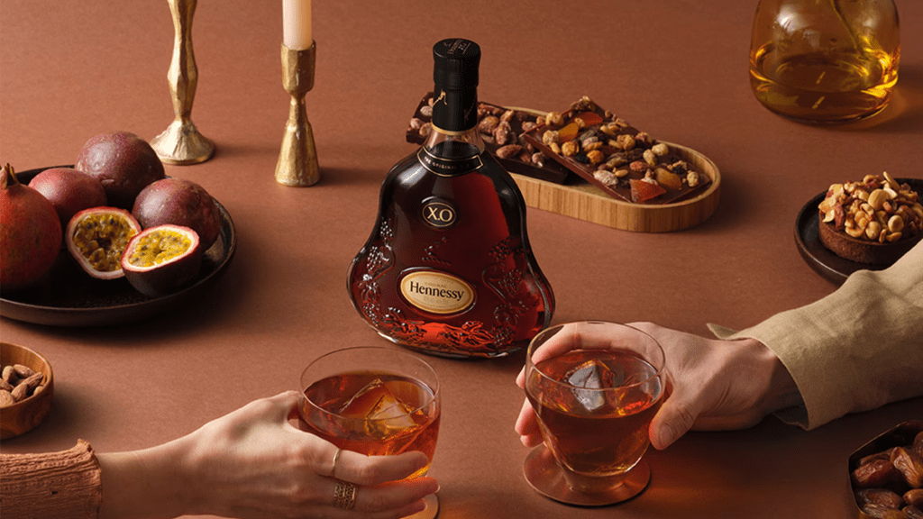 A tablescape in soft, warm colour tones. At the centre is a bottle of Hennessy XO and two hands holding cocktail glasses with cognac inside them. Scattered around the table are various adornments like candlesticks, a bowl of fruit, and more.