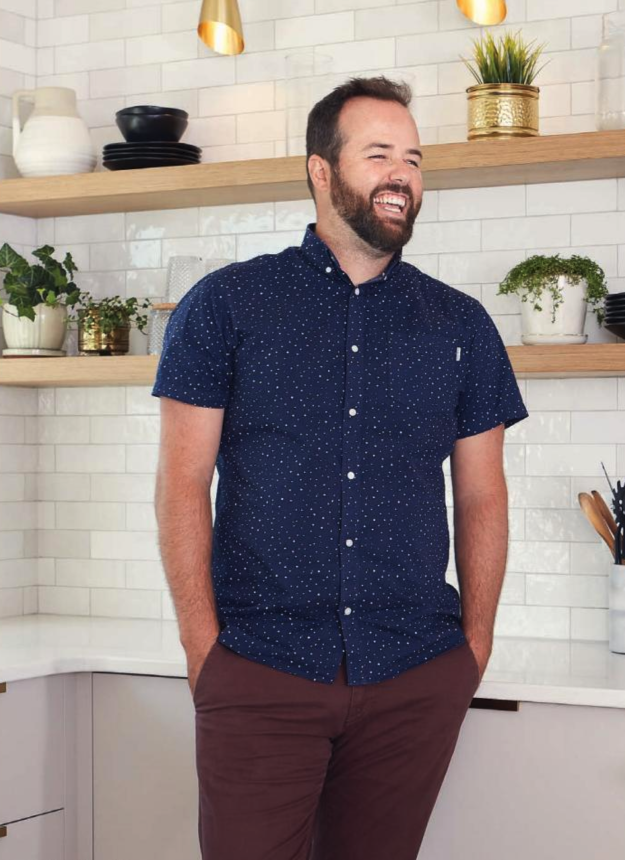 Andrew Hibbs wearing a short-sleeved blue button up. His hands are in the pockets of maroon pants. Backdrop is a kitchen corner with wooden shelving.