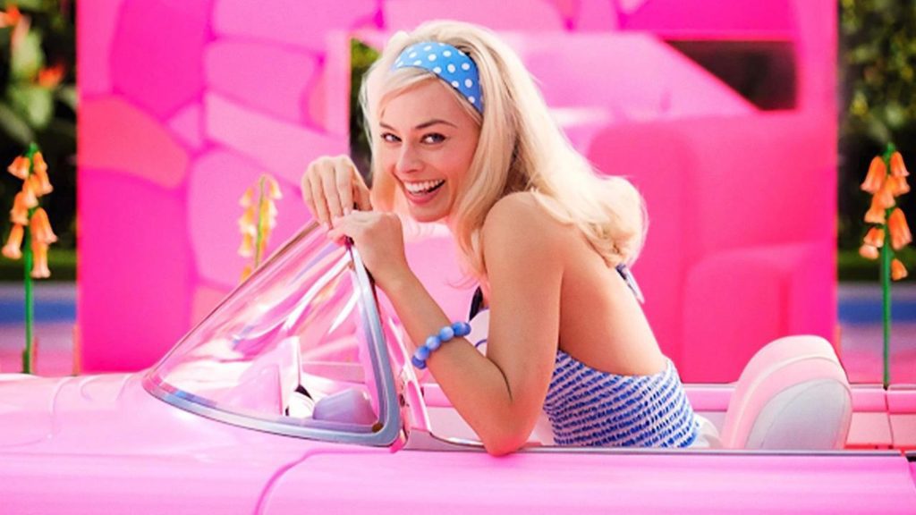 Margot Robbie smiling and sitting in the driver's seat of a bright pink convertible car. She is wearing a blue polka dot headband with a matching dress and bracelet.