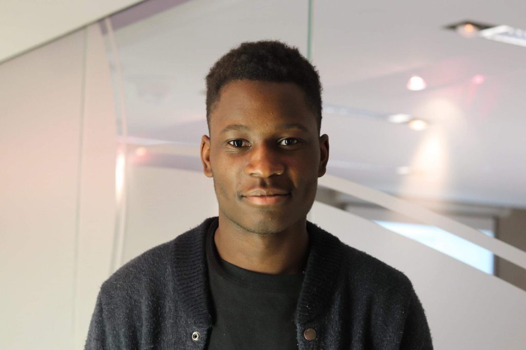 Man wearing a black t-shirt and jacket smiling into the camera. He is centered with office lighting in the background.