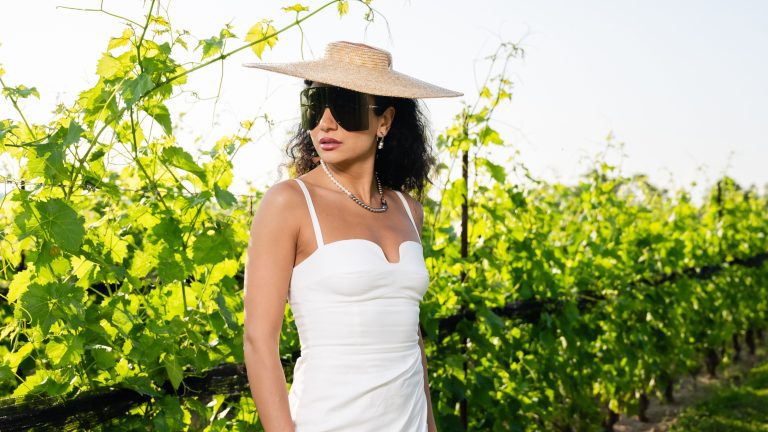 Dorian Who wearing a white dress, oversized sunglasses and a straw broad-brimmed hat against a backdrop of vineyards