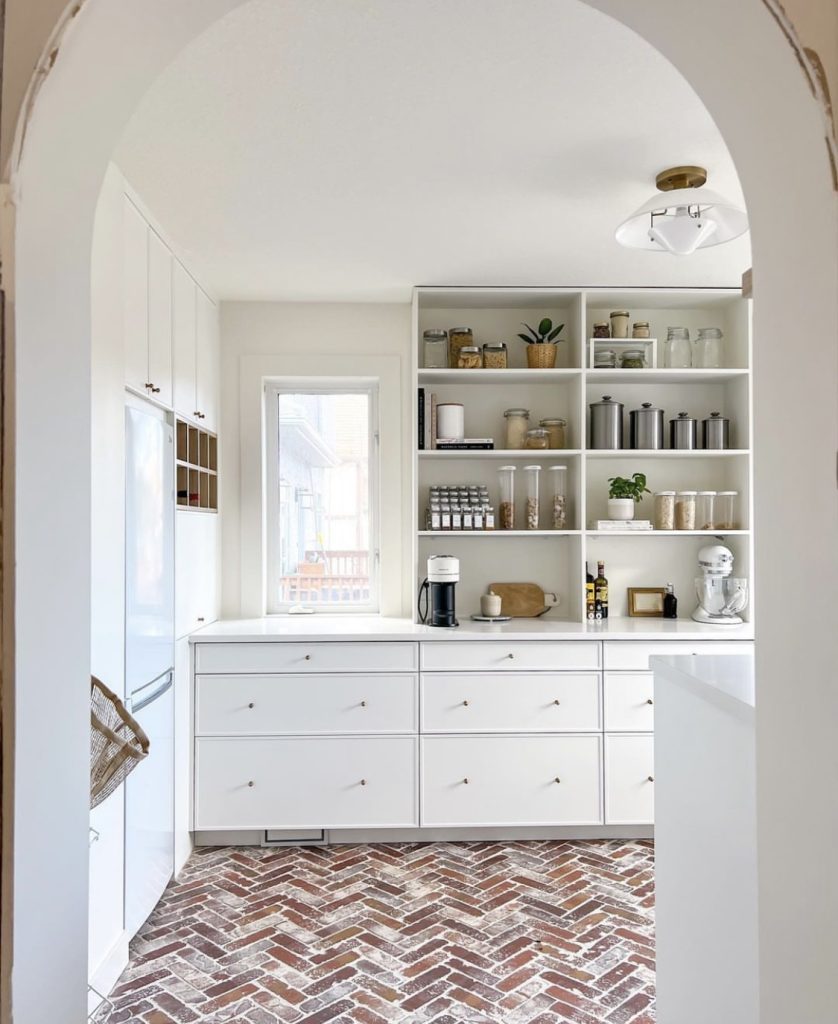 Kitchen arched entryway. There is a herringbone tiled floor in various stones. Featured alongside it is a kitchen storage system in white with gold hardware and shelving above it that goes to the ceiling with various cannisters and household items.