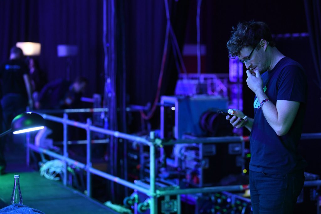 Paddy Cosgrave looking pensively down at his phone wearing a black t-shirt. He is in a dark room, presumably backstage at the Collision conference.