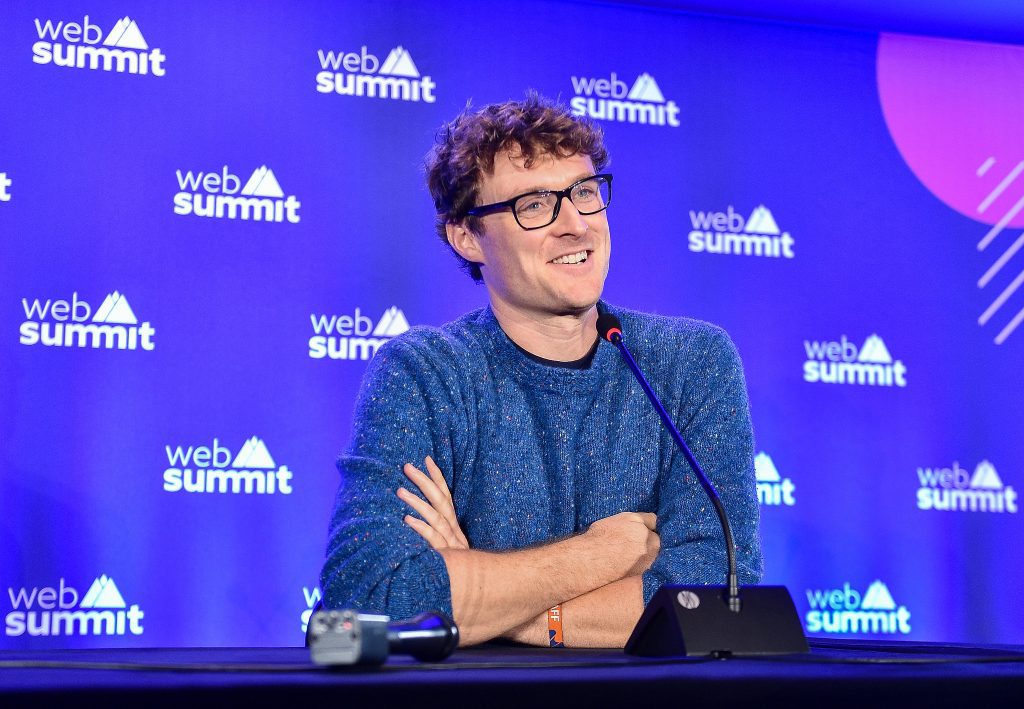 Paddy Cosgrave wearing blue sweater and sitting at a press conference table. His arms are folded and he is smiling against a backdrop with the Web Summit logo.