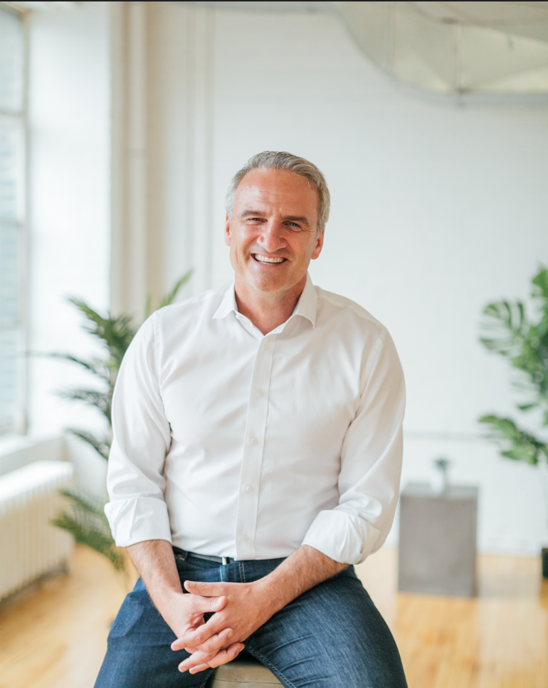 Michael Garrity, CEO of the tech company Financeit, sitting on a stool in a white room with some plants in the background. He is wearing blue jeans, a white, button down shirt, and he is smiling.