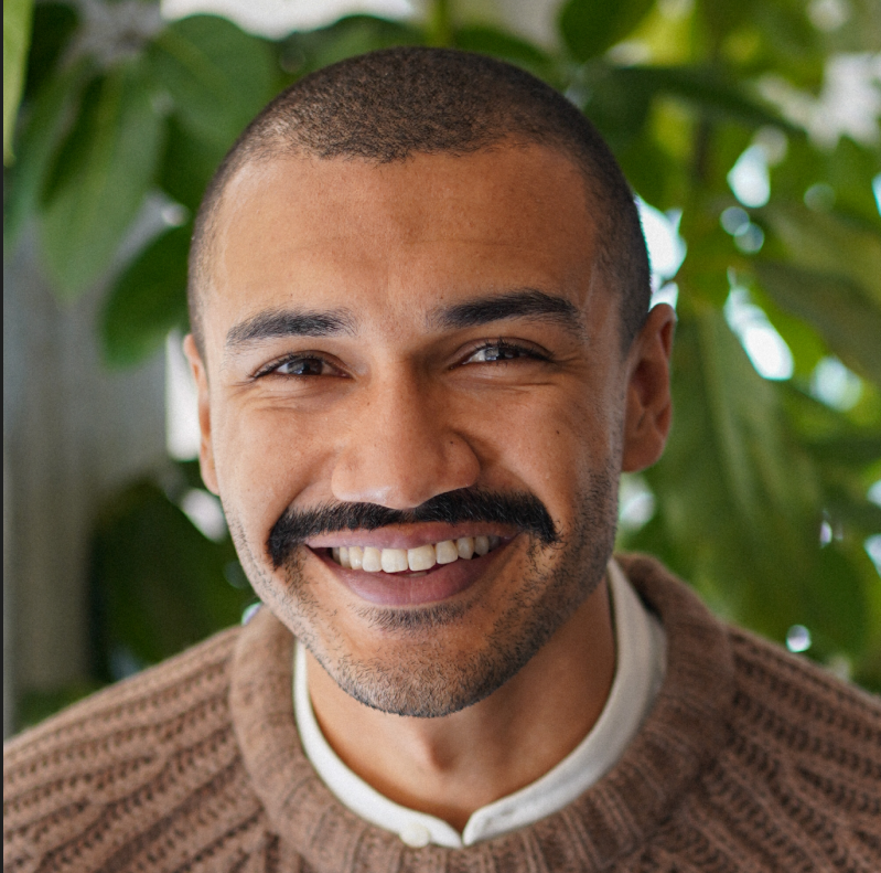 Close up photo of Brandon Goode, co-founder of Outro, the antidepressant tapering platform. He is wearing a brown sweater with a white shirt underneath and he is smiling. There are green leaves behind him.