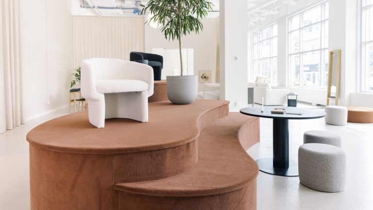 Bright showroom with a terra cotta coloured two-step platform. On top of it is a plus white seat and a large plant in a concrete planter.