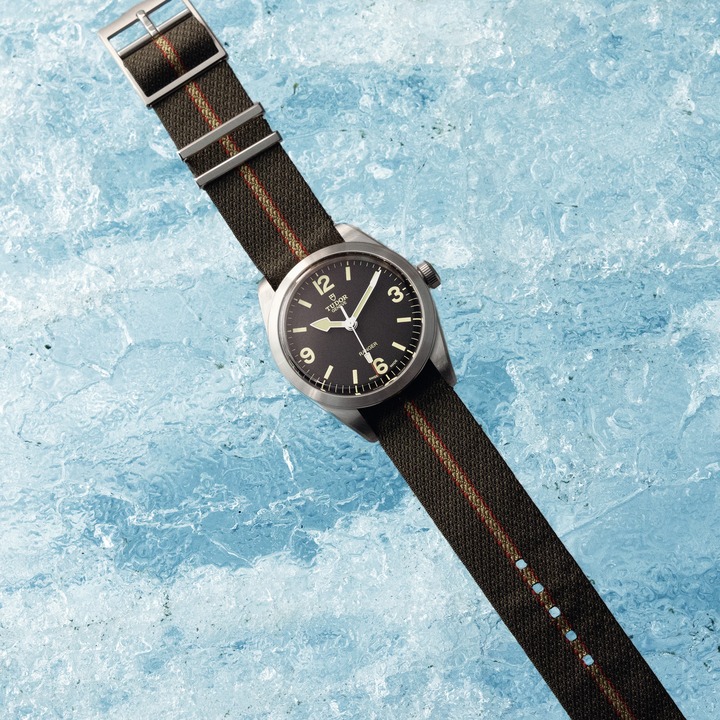 On a sheet of blue cracked ice is a TUDOR Ranger watch laid flat. The strap is a green canvas with a light brown stripe going down the middle. The watch has a black face and steel dial.