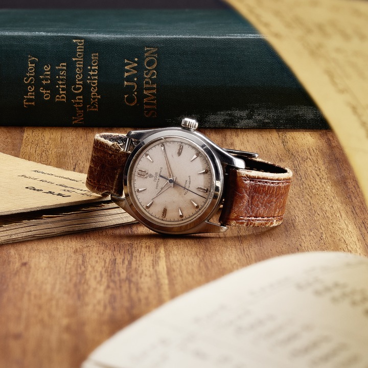 Vintage TUDOR watch lying on its side. The face is aged and the brown leather straps are aged. It is sitting against a journal on a wood table. In the background is a green book spine.