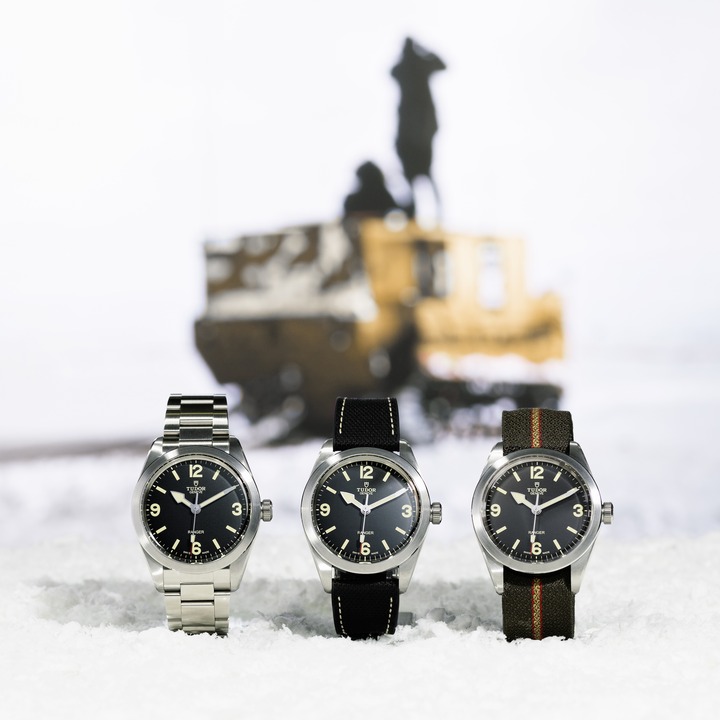 Three TUDOR watches set on ice. One has a steel bracelet, the middle watch has a black strap, and the right watch has a khaki green strap. All of them have black faces. In the background is a snow tank vehicle.