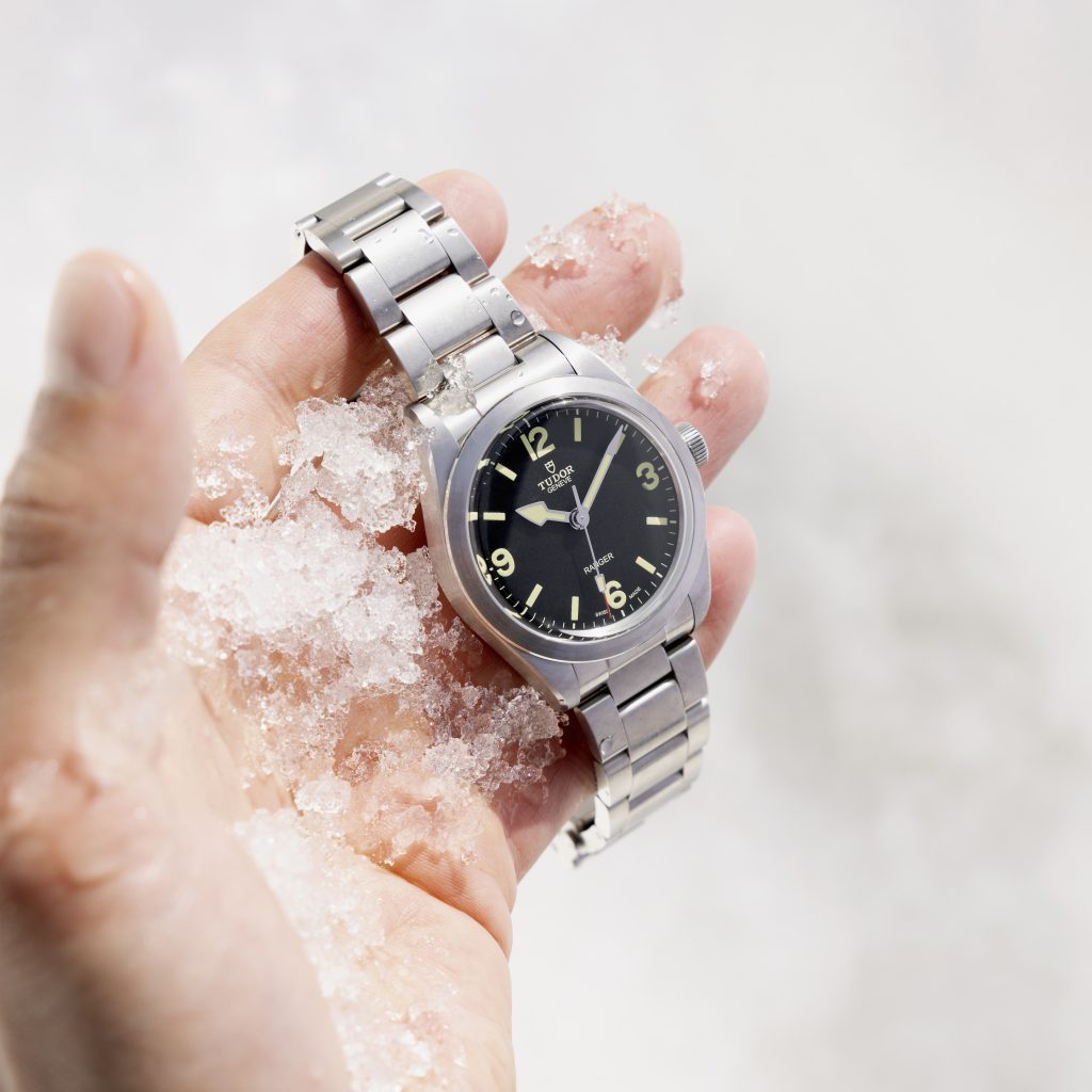 A hand is holding a steel TUDOR Ranger watch with a black face. The hand has bits of ice on it and the background is seemingly a blurred image of ice.