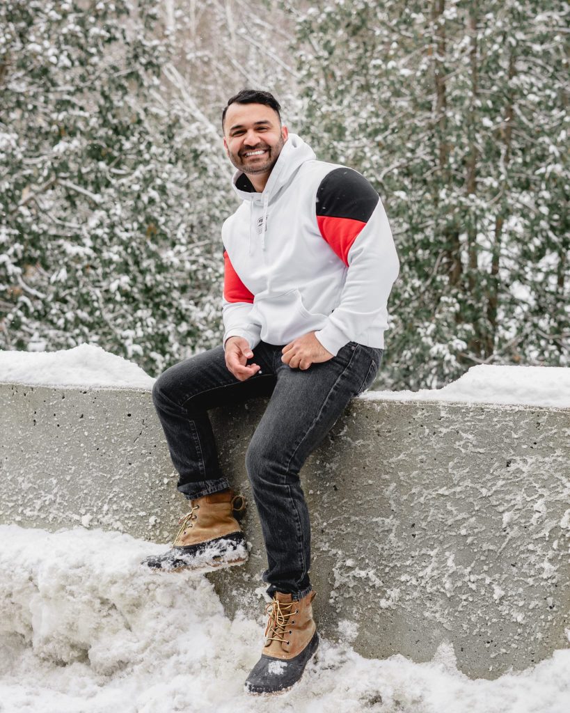 Photo of Sunny sitting outside in winter. He is smiling, and wearing a grey sweater. There are some trees behind him with snow on them.