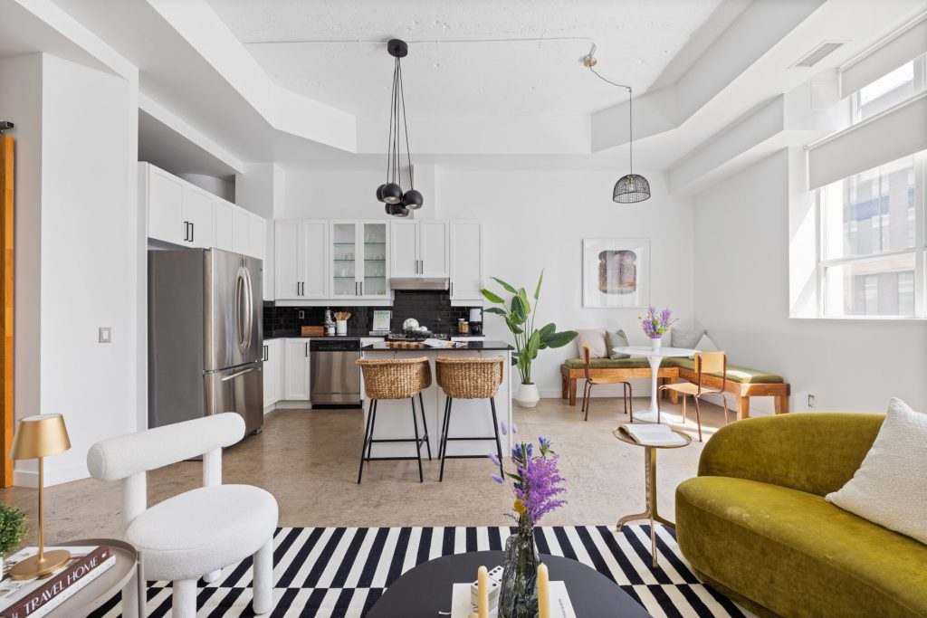 Industrial light-filled loft with a cozy kitchen with white cabinetry and a breakfast bar. There is a green sofa situated in front of a black coffee table and white arm chair. They are on top of a black and white rug.