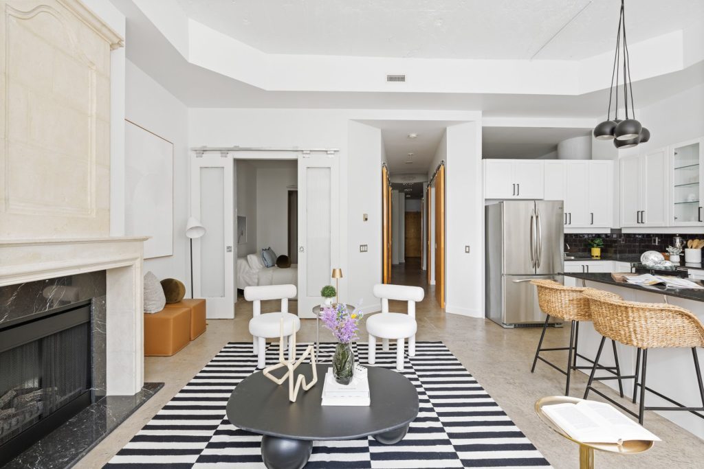 Industrial light-filled loft with a cozy kitchen with a marble fireplace mantle. There is a black coffee table and two white arm chairs in front of a bedroom and hallway. They are on top of a black and white rug.