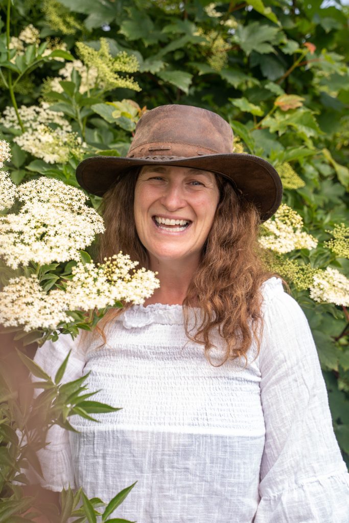 Geraldine Kavanagh smiling and wearing a brown brimmed hat and white blouse. She is against foliage of a bush with flowers.
