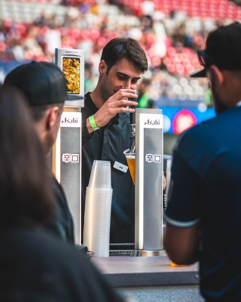 Man pouring Asahi beer from a draught machine at a soccer game.