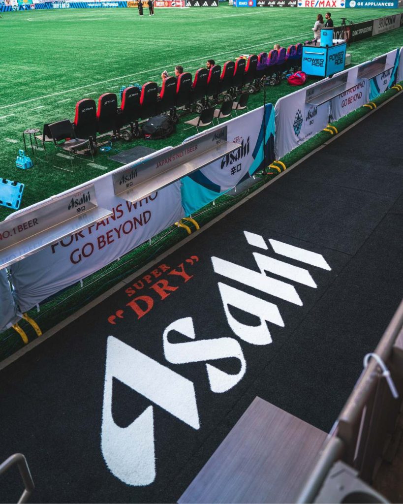 Black carpet with large Asahi beer branding. It is overlooking a row of seats that look out onto a soccer field.