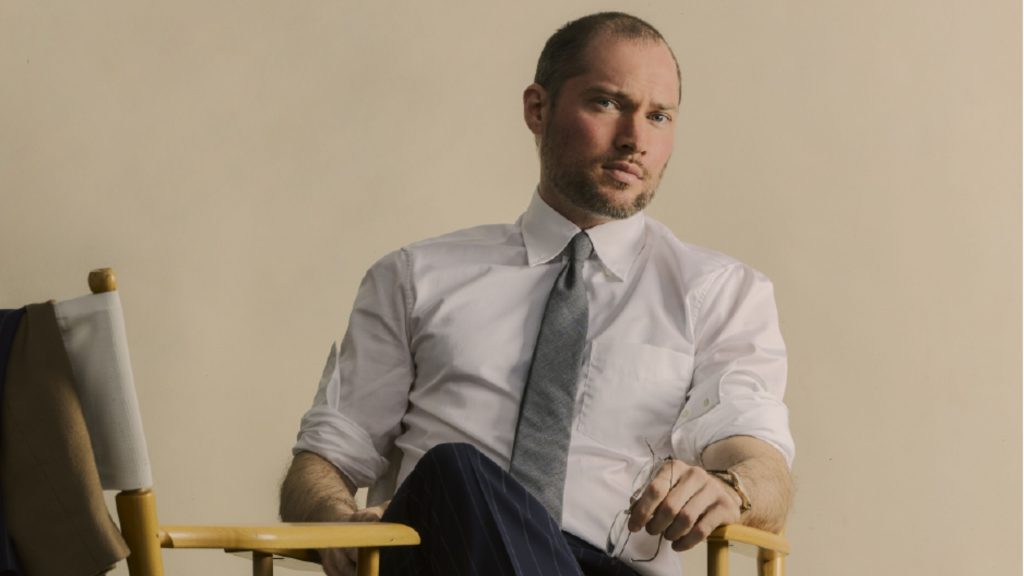 Oliver El-Khatib looking stoic into the camera. He is wearing a white button-up shirt with a grey tie and navy blue pants. He is seated in a director's chair against a beige background.