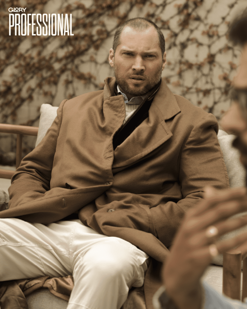 Oliver El-Khatib seated wearing a camel overcoat and white pants. Behind him are some dried up ivy vines.