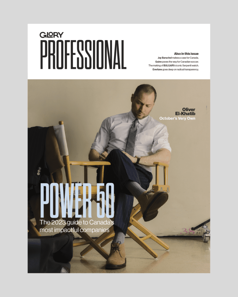 Magazine cover with Oliver El-Khatib, founder of October's Very Own, sitting in a director's chair. He is wearing a white button up with a grey tie and blue pants. Legs are crossed and he is looking down.