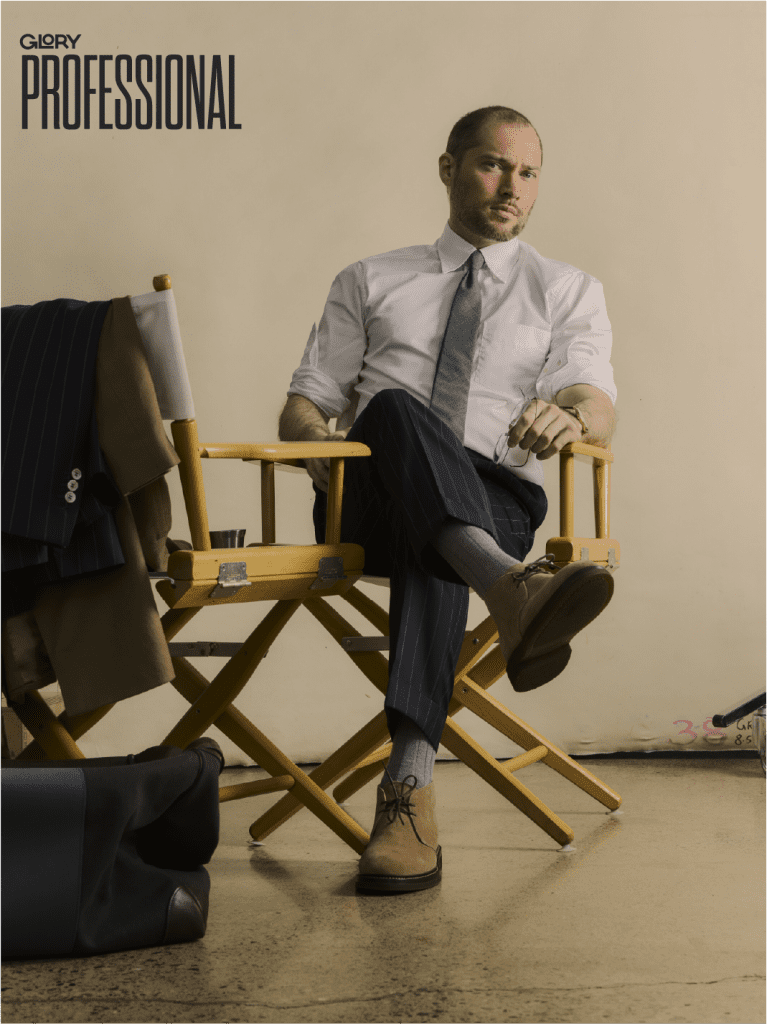 Oliver El-Khatib looking stoic into the camera. He is wearing a white button-up shirt with a grey tie and navy blue pants. He is seated in a director's chair against a beige background.