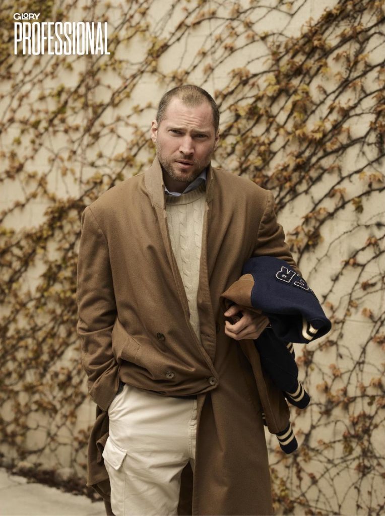 Oliver El-Khatib standing wearing a camel overcoat and white pants. He is also holding a varsity jacket in the crook of his arm. Behind him are some dried up ivy vines.