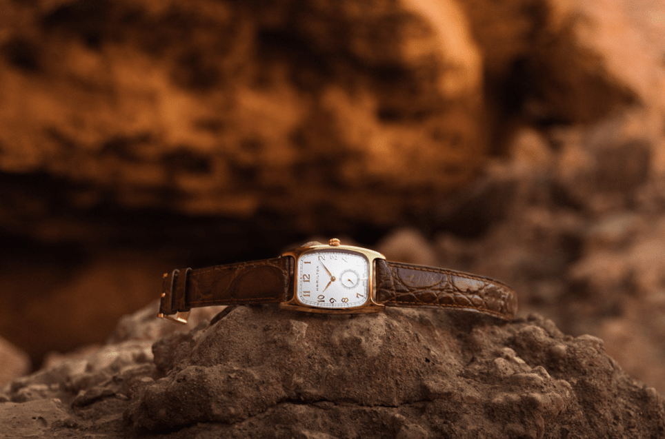Timepiece on its side. It has a rectangular face with a brown leather strap and is on top of a rock with more rocks in the background out of focus.