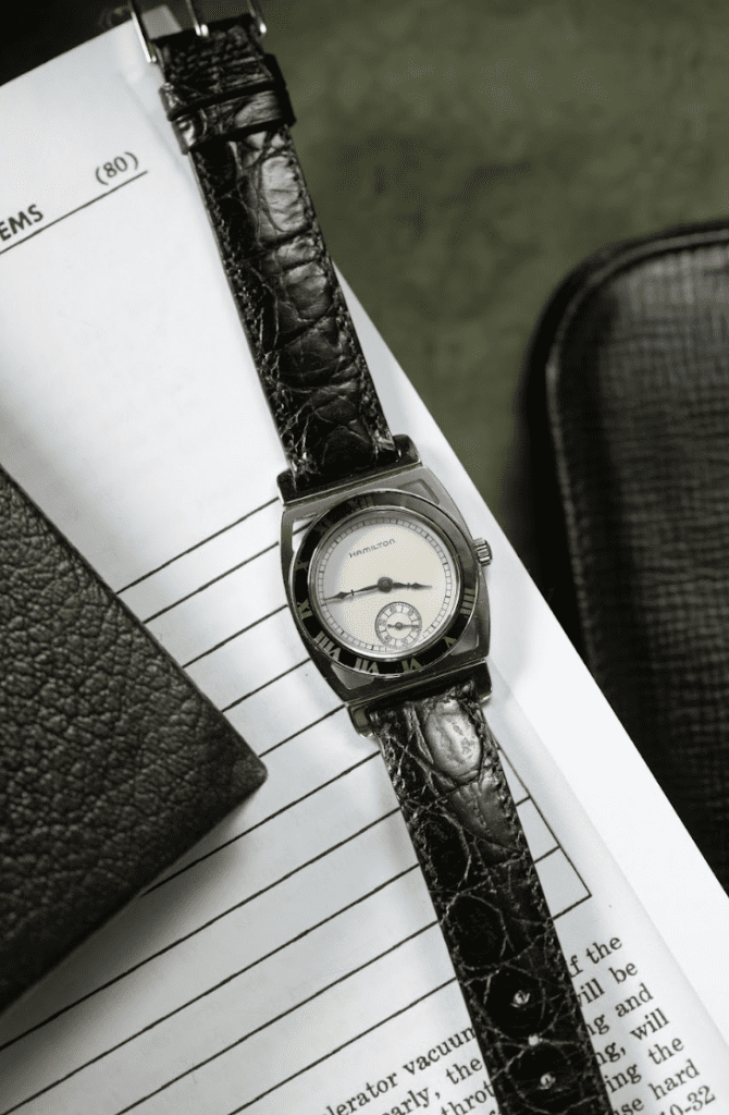 Hamilton watch with a black strap and silver dial against a piece of lined paper and the corner of a leather notebook.