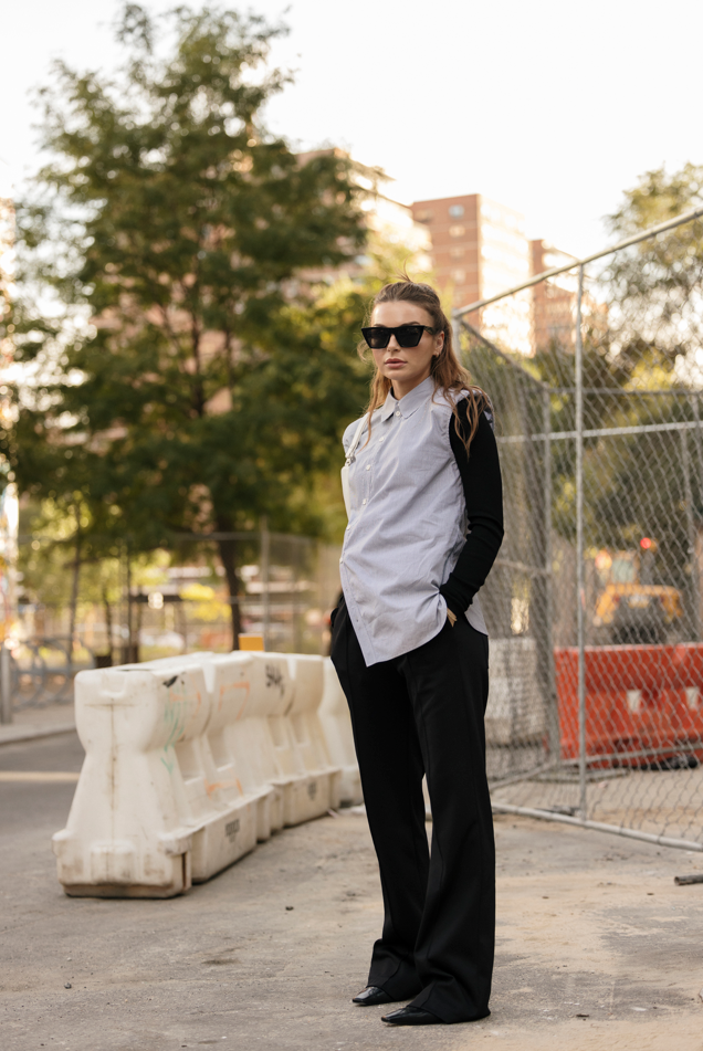 Photo of Allegra Shaw standing outside wearing black pants and a grey button up shirt. She is also wearing dark sunglasses