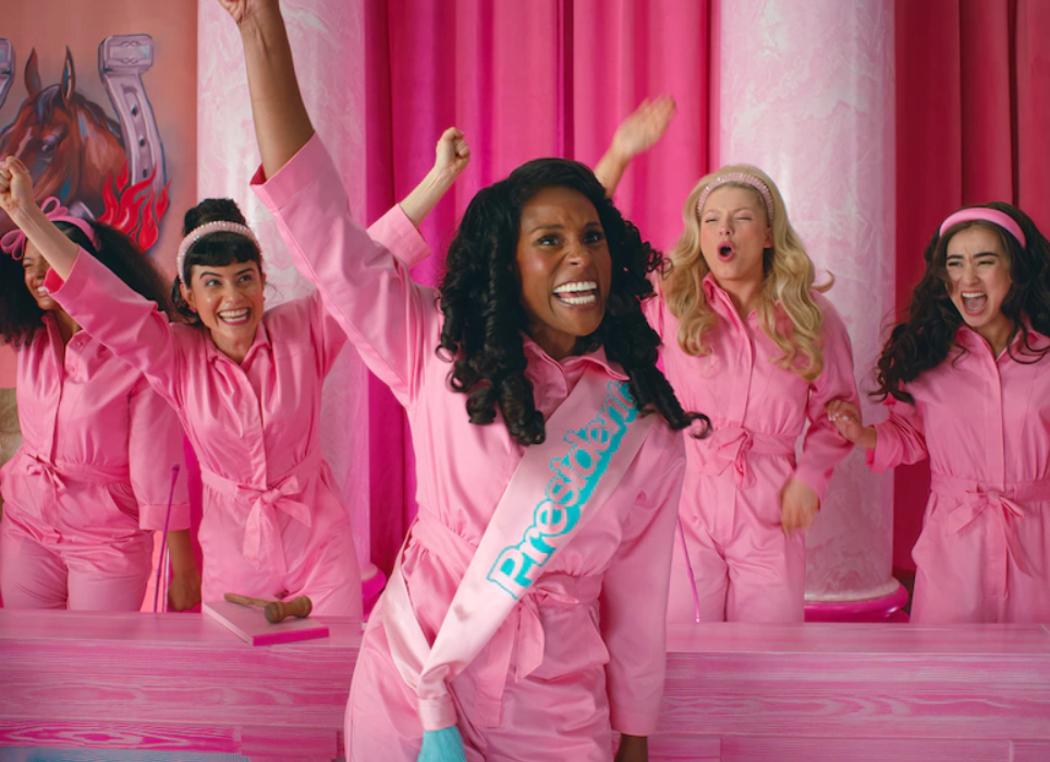 The president of Barbie Land smiling with her arm in the air. She is dressed in all pink and is wearing a sash that says "President" in blue writing. There are other Barbie's standing behind her also wearing pink and smiling with their hands up in the air.