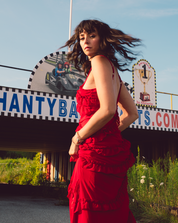 Photo of a women in a red dress. Her body is facing a go carting sign but she is turning her head towards the camera.