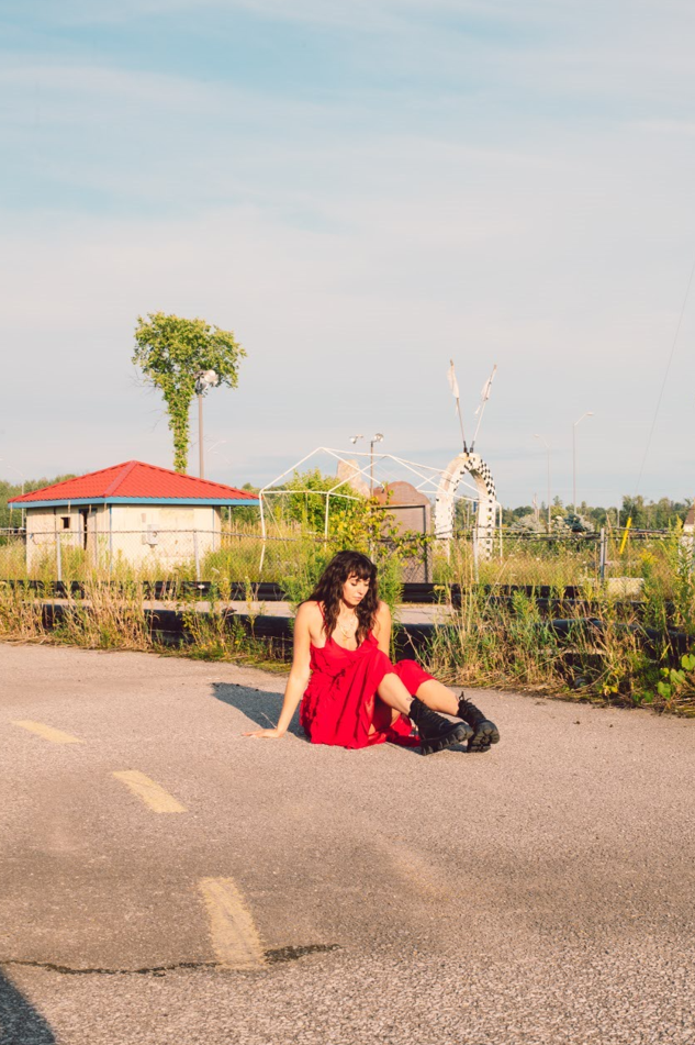 Image of Briony sitting in the middle of the road. She is wearing a red dress with black boots, and she is looking down.