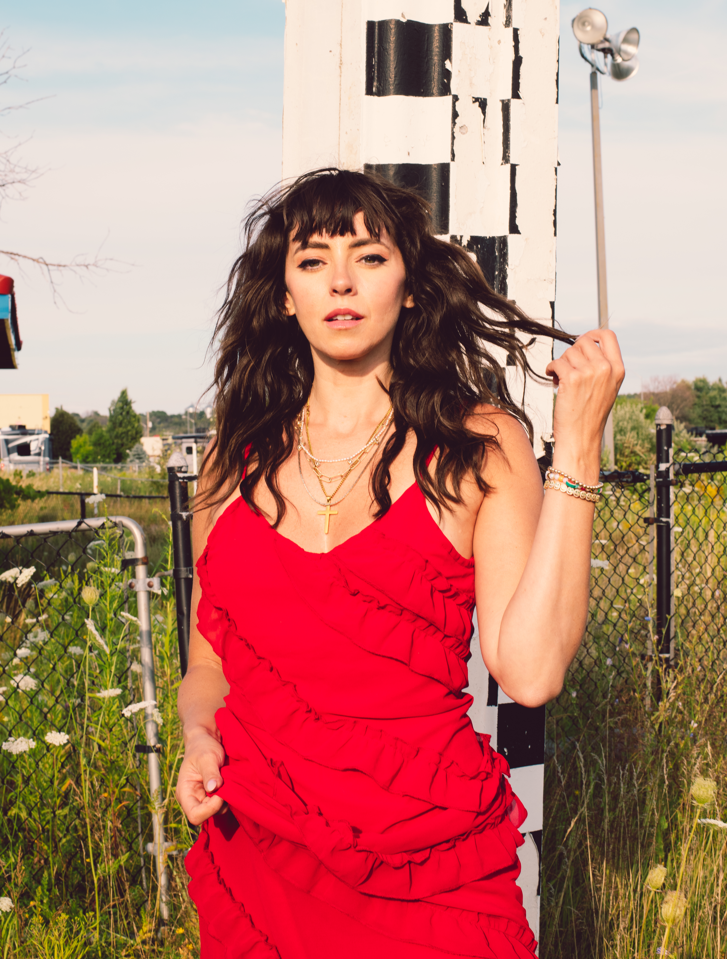 Photo of Briony looking at the camera. She is wearing a red dress and twirling a bit of her hair in one of her hands. There is a fence behind her with a grassy field.