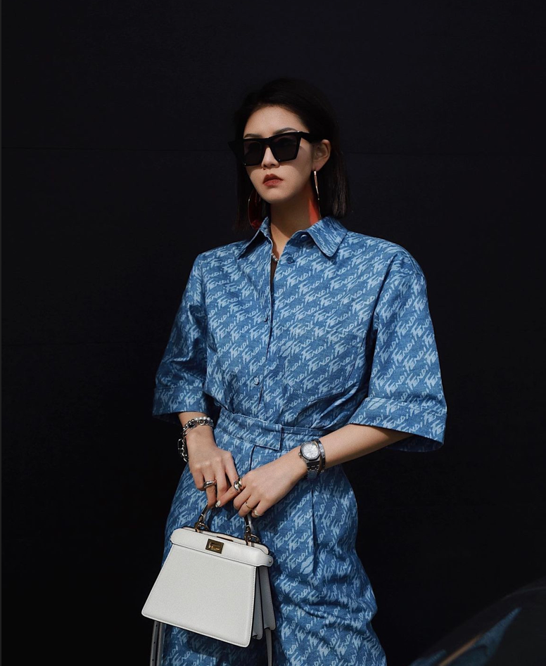 Photo of Yuki Zhao in front of a black background wearing a blue overcoat with a white pattern on it. She is holding a white handback in front of her and she is wearing black sunglasses.
