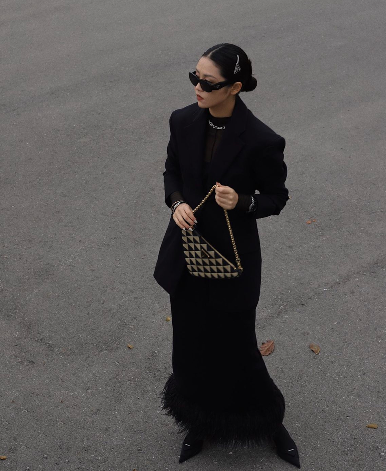 Photo of women wearing all black, holding a black bag, and wearing black sunglasses. She is standing on some pavement and looking off the side.