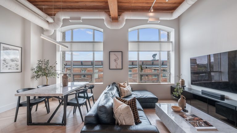 Modern urban loft with wood beam ceilings, tall windows and chevron floors. There is a black couch in front of a TV with a long dining table and chairs.